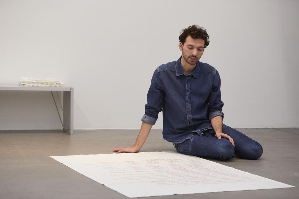 artist sits on floor in front of large fabric with texts on it and reads from it